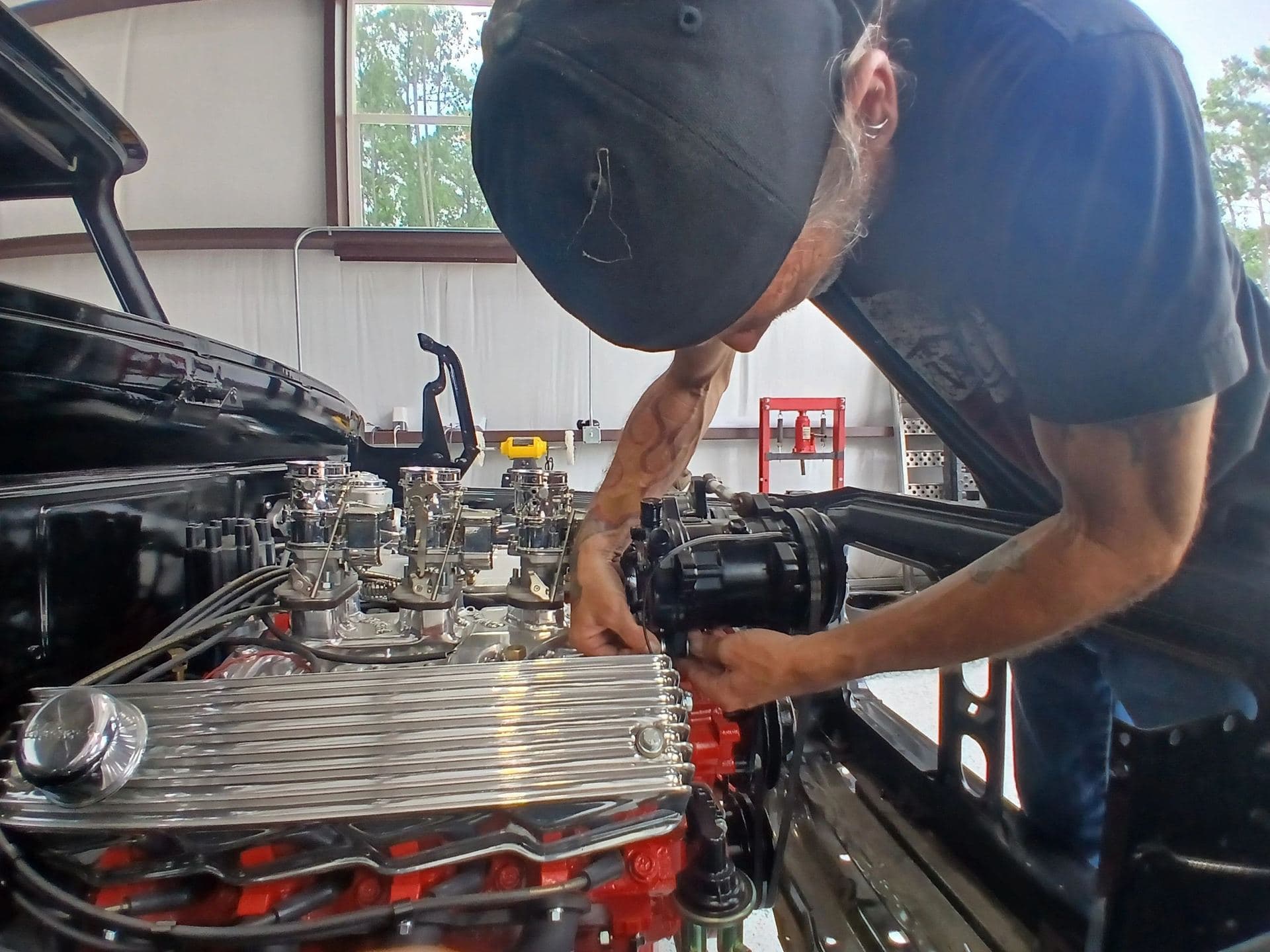 Mechanic working on a red tri-power engine in a black hot rod at Tha Shop